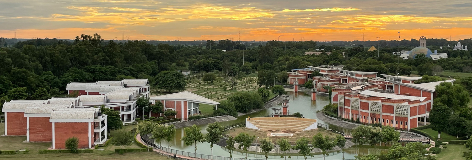 Aerial view of a campus with modern red-brick buildings, lush green trees, a central circular amphitheater, and a pond, under a cloudy orange sunset sky