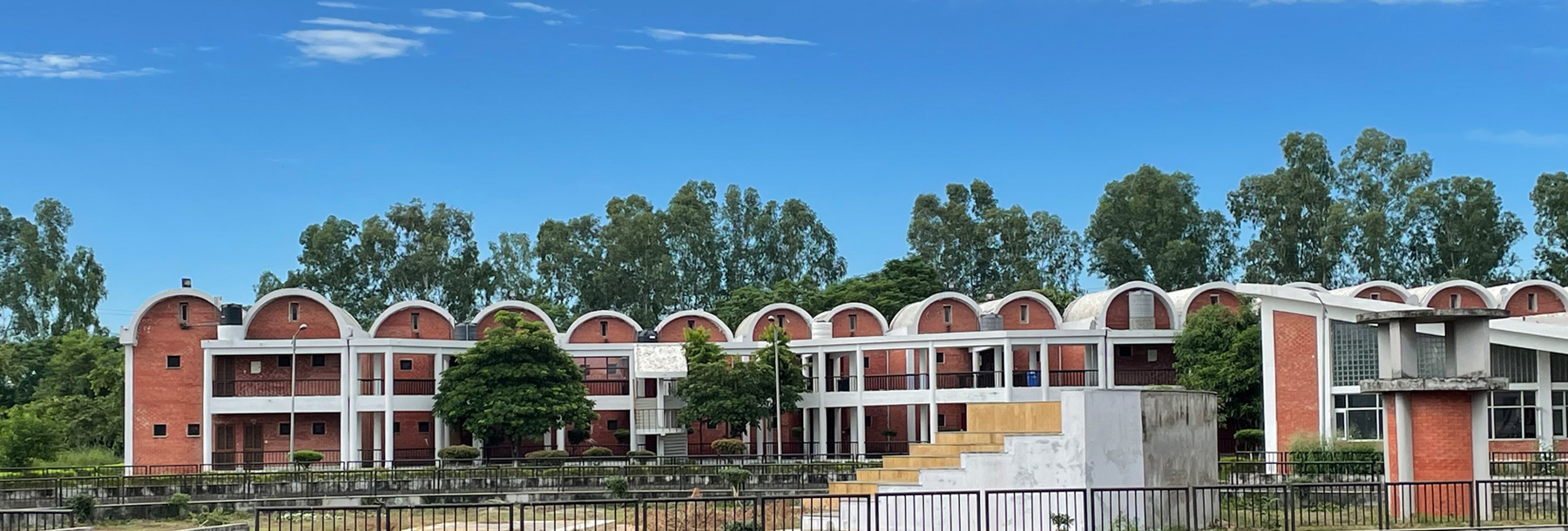 A row of red brick buildings with arched white roofs surrounded by trees under a blue sky, with a concrete structure and steps in the foreground