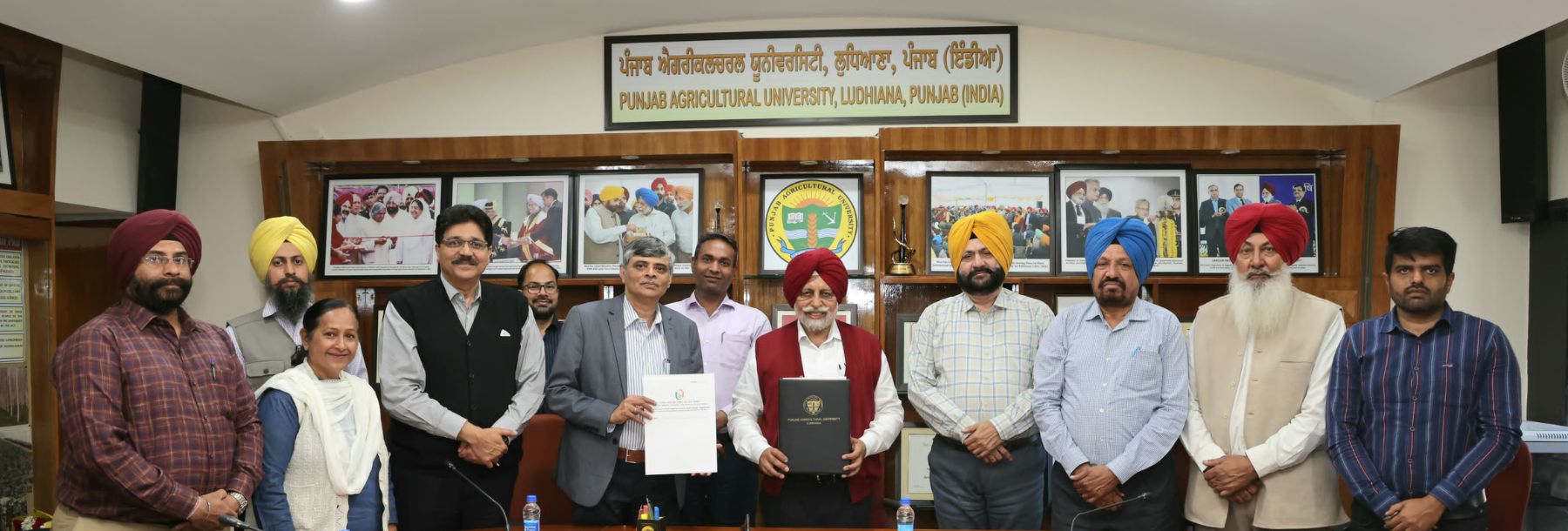 A group of people, including men and women, many wearing turbans, stand and pose for a photo in a conference room at Punjab Agricultural University, holding documents and smiling at the camera.