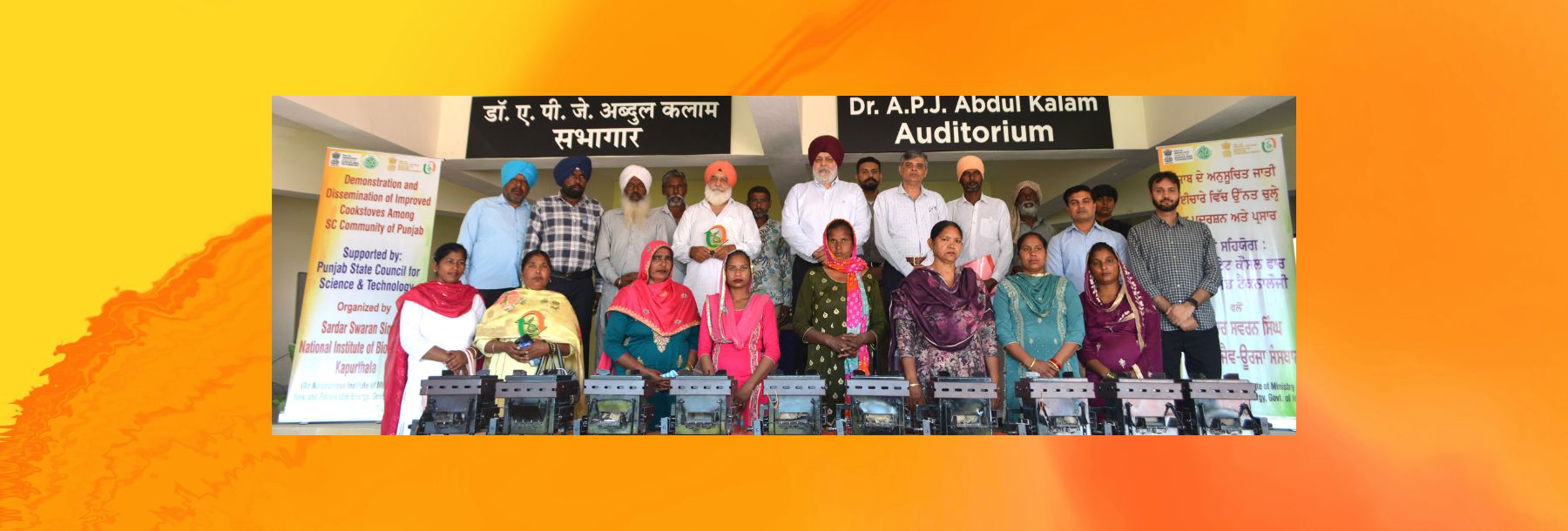 A group of men and women, some in traditional Indian attire, stand and sit posing for a photo on a stage labeled Dr. A.P.J. Abdul Kalam Auditorium with banners and sewing machines in front of them