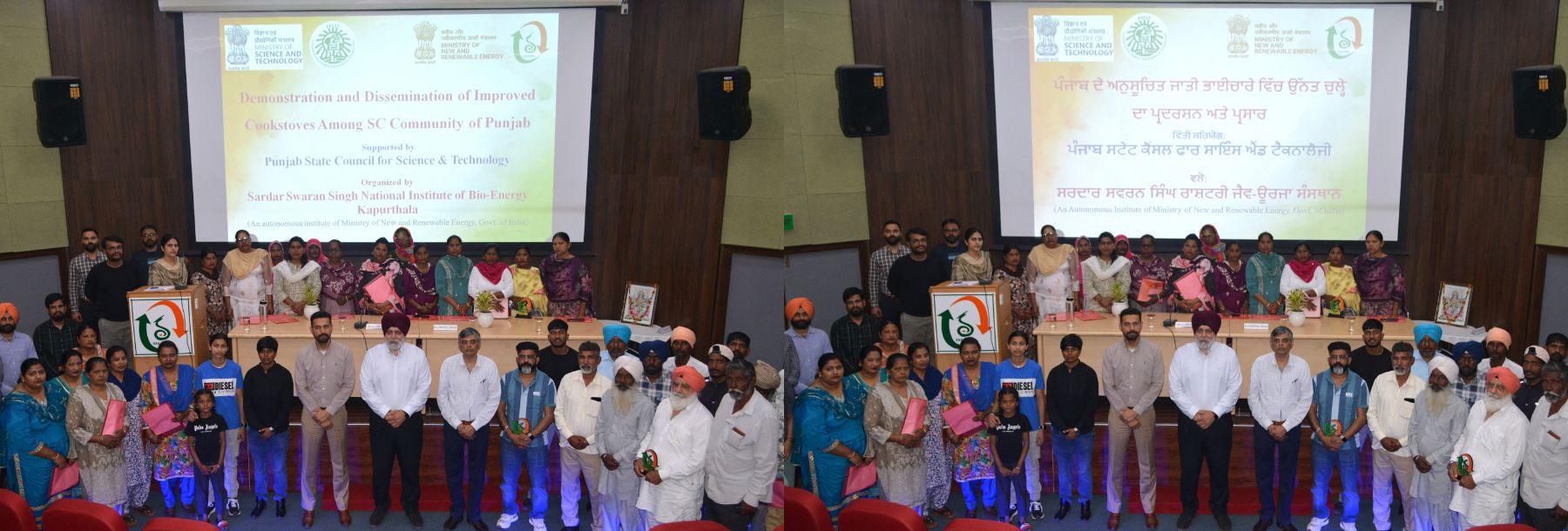 A large group of people, including men and women, stand and sit on a stage in front of a banner with text in English and Punjabi at a formal event in an auditorium. Tables, microphones, and chairs are visible.